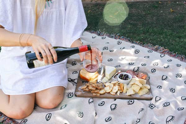 Woman pouring red wine into a stemless wine glass on a blanket next to a charcuterie board.