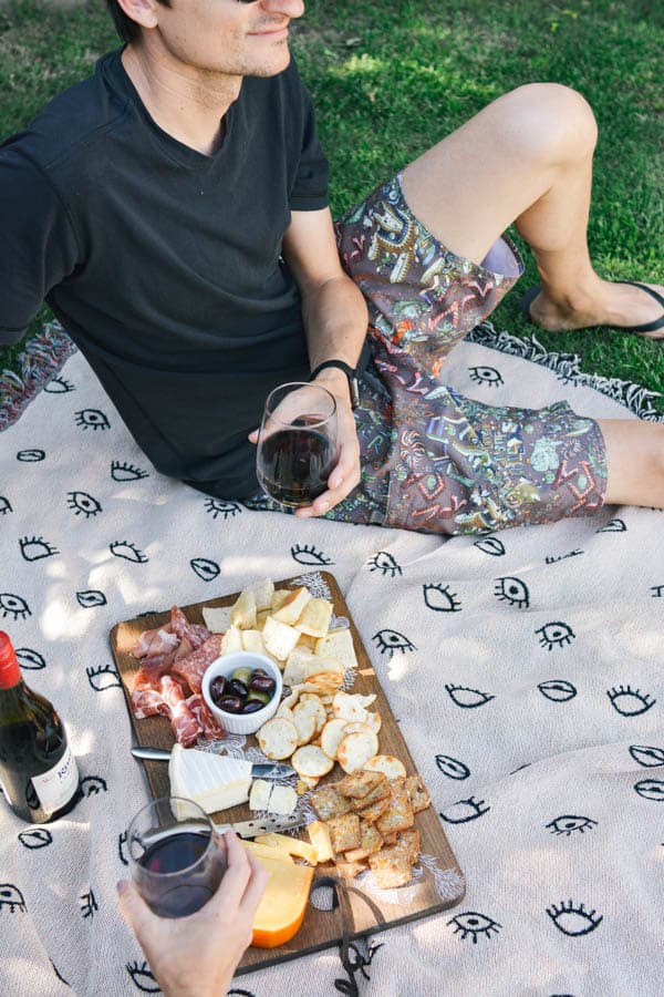 A man sitting on a picnic blanket holding a glass of red wine next to a charcuterie board.