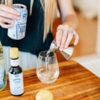 Woman pouring club soda into a glass of wine to make a white wine spritzer.