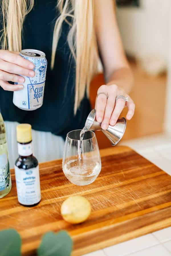 Woman adding club soda from a jigger into a stemless wine glass on the counter. 