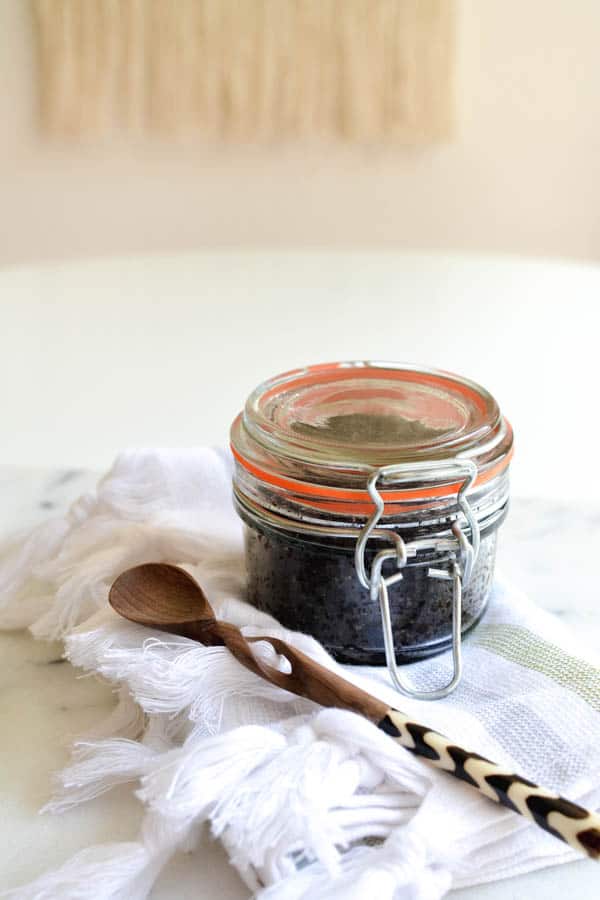 Close up image of a glass jar filled with DIY coffee scrub on top of a towel next to a wooden spoon.