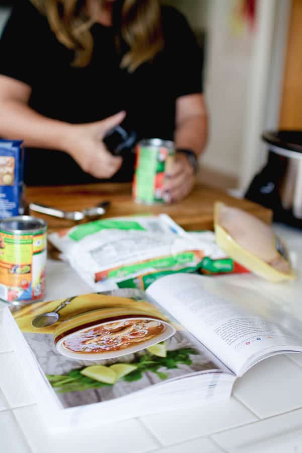 A cookbook on the counter with a woman opening a can of tomatoes in the background.