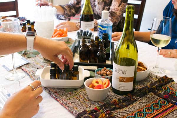 Women reaching for a bottle of essential oils on a table with snacks and bottles of wine on the table. 