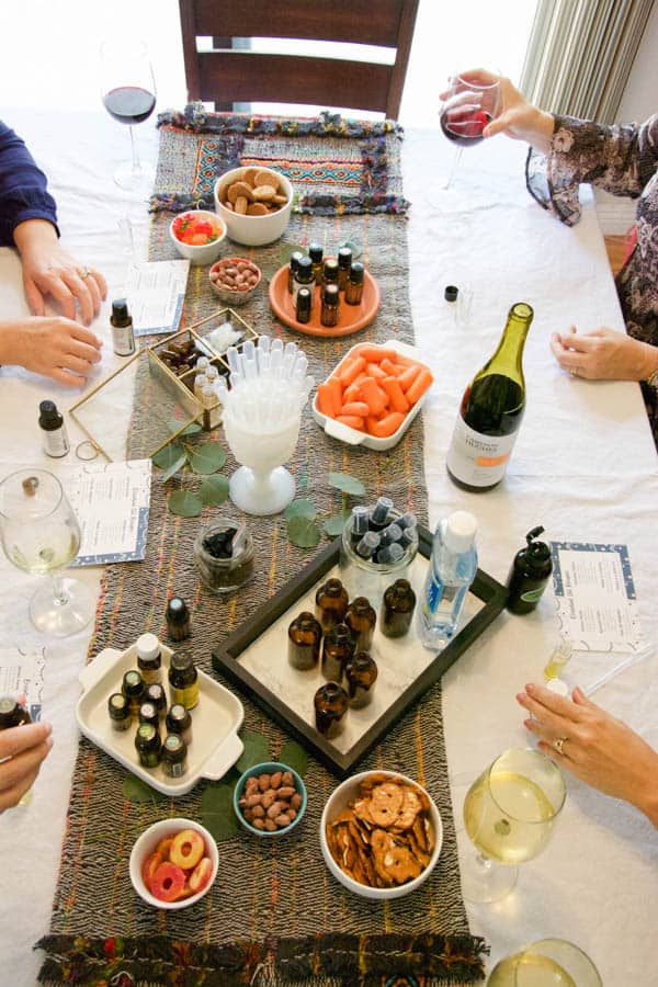 Friends enjoy wine around a dining table covered with snacks and supplies to make holiday essential oil blends. 