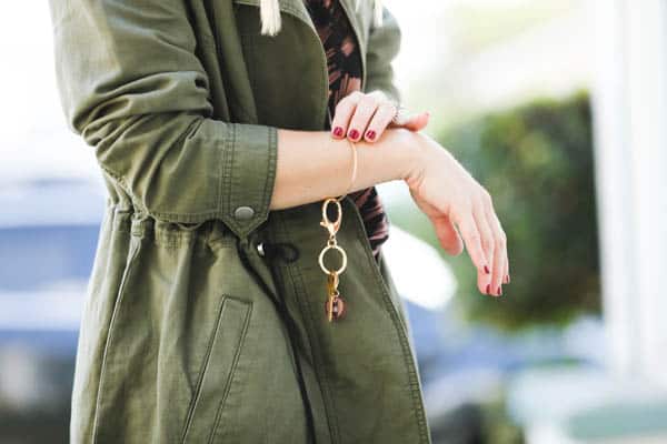 Woman wearing green jacket wearing a gold bracelet with keys on it and touching the bracelet with her other hand.