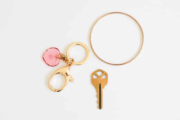 A gold keyring attached to a clasp and pink heart next to a key and gold bracelet laying flat on a white table.