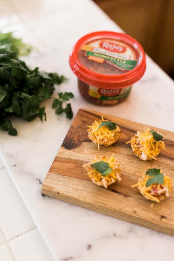 A small wood tray on the counter with Chicken Taco cups in phyllo cups next to a container of salsa. 