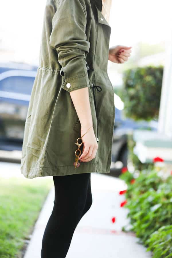 Girl in black pants and green jacket with her arm at her side wearing a gold bracelet with keys on it.