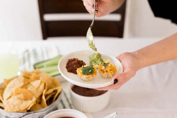 Woman holding a small white appetizer place with Chicken Taco Cups and salsa and adding guacamole from a spoon. 