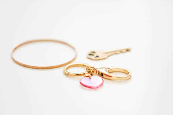 Gold bangle bracelet, a key, and a keychain ring attached to a clasp with a pink heart sitting on a white table.