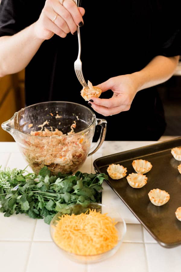 Woman making taco cups with shredded chicken, cheese, cilantro and phyllo cups. 