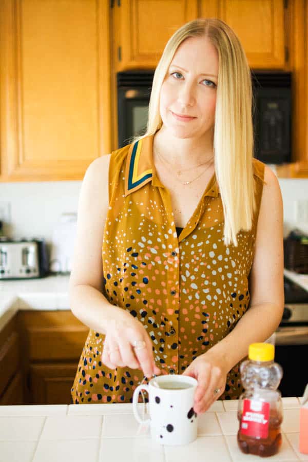 Blonde woman in mustard colored sleeveless blouse smiling and making tea.