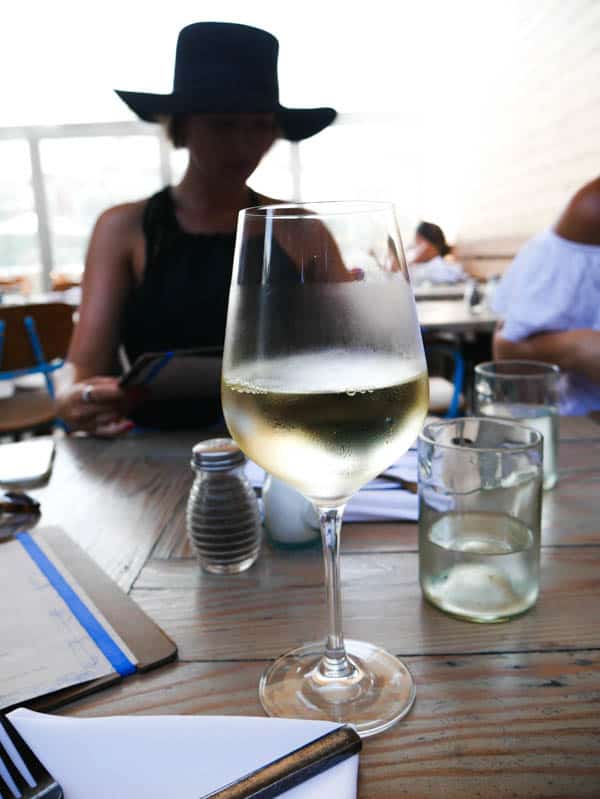 Close up of white wine in a wine glass on a table at a restaurant with the silhouette of a girl in a hat in the background.