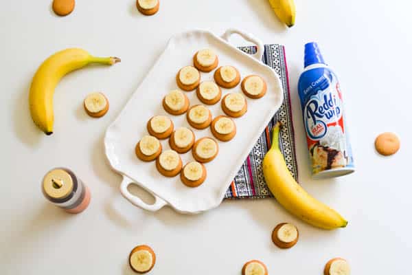 A tray of Nilla Wafers and sliced bananas on a table next to a can of whipped cream, a bottle of caramel and bananas. 
