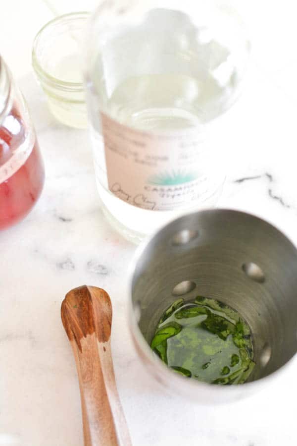 Cocktail shaker with muddled basil and lime juice next to other ingredients on a cutting board. 