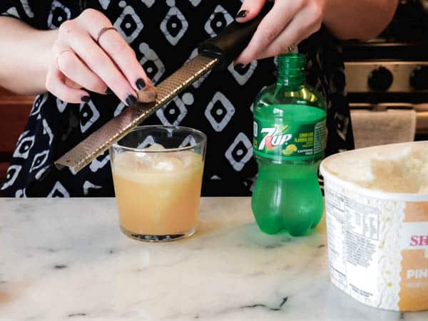 Woman grating fresh nutmeg on top of a cocktail in a short glass on a counter. 