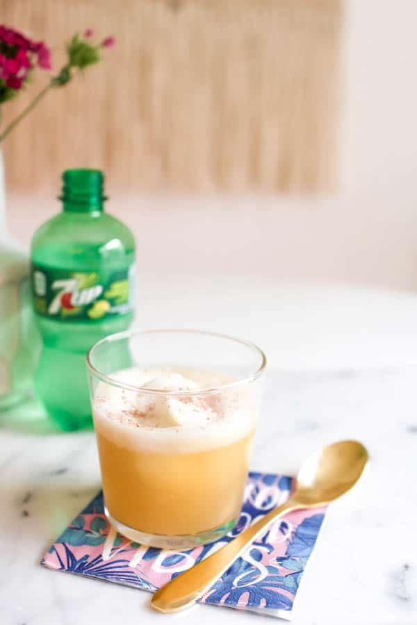 A pineapple float cocktail in a glass topped with fresh nutmeg on a table next to a spoon and a bottle of 7UP.