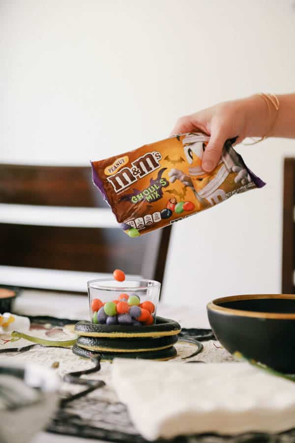 Woman adding Halloween peanut M&Ms into a small glass wrapped with a plastic snake. 