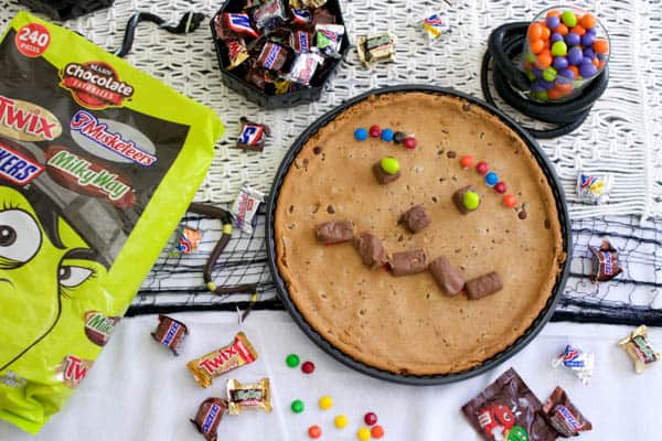 Monster face made out of candy pieces on a large cookie on a Halloween party table with a bag of candy for a Halloween playdate.