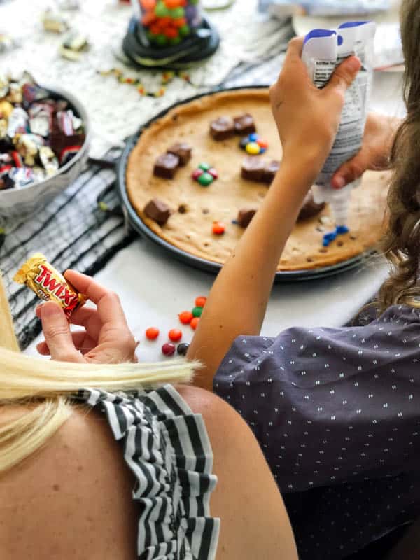 Over a woman's shoulder of her holding a small candy bar while a kid decorates a giant cookie. 