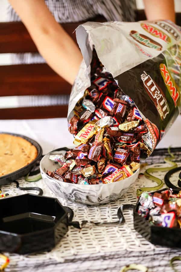 Woman pouring fun-sized candy bars into a serving bowl on a Halloween party table. 