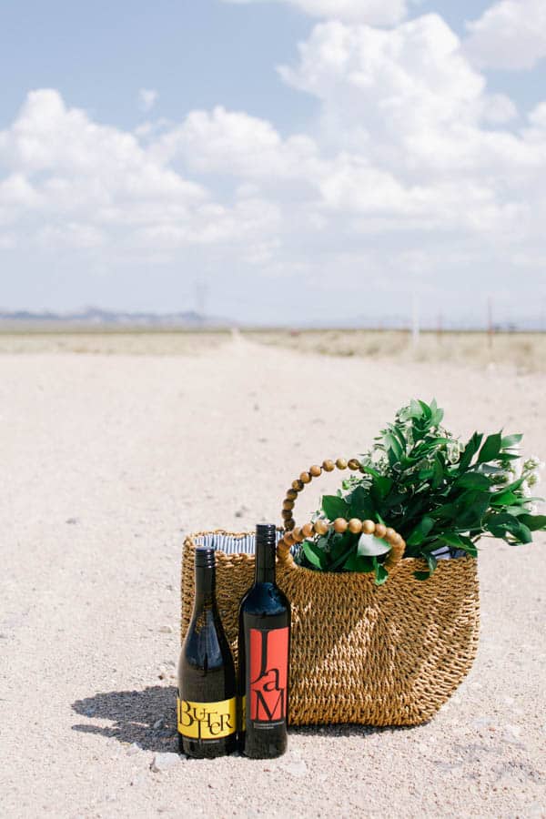 An oversized tote bag with flowers sticking out and wine bottles next to it on an open dirt road. 