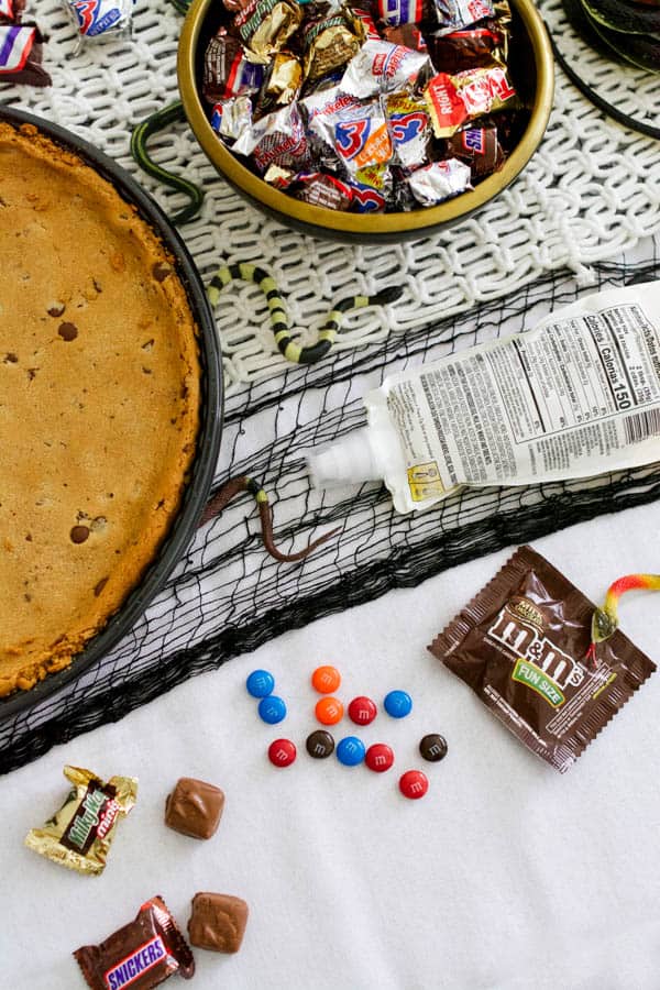 A giant cookie next to a bowl of candy and an icing bag on a Halloween party table. 