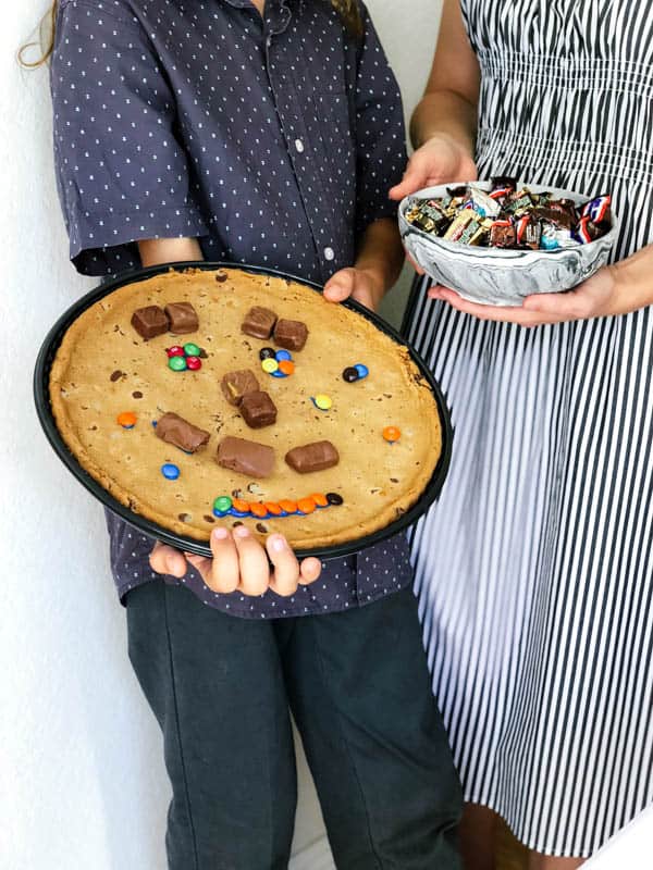 Kid holding an extra large cookie with a face made of candy and a mom holding a bowl of candy.