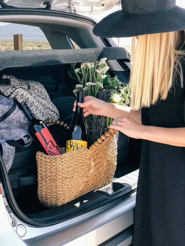 A woman placing a bottle of wine in a tote bag in the back of a car before setting out on a road trip. 