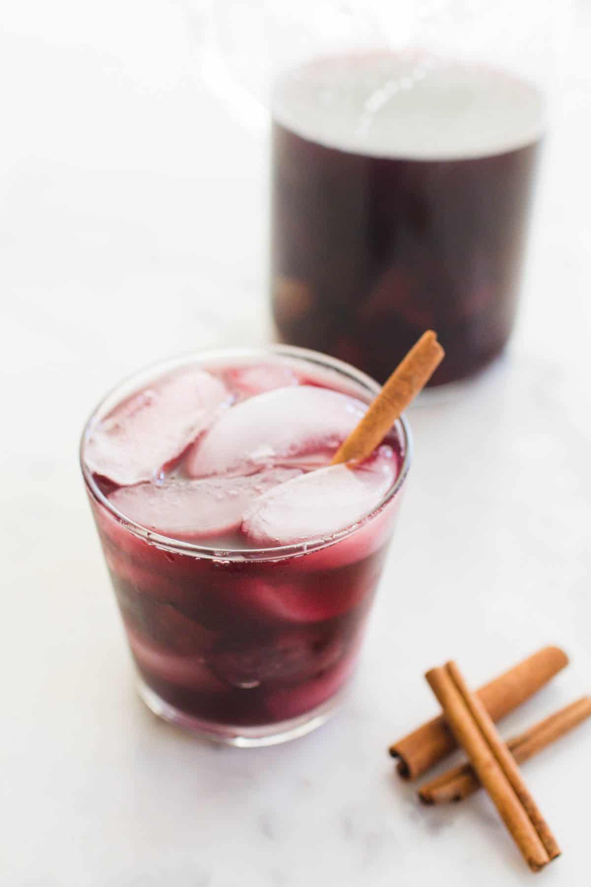 Glass of red wine fall sangria on a table near the pitcher and cinnamon sticks.