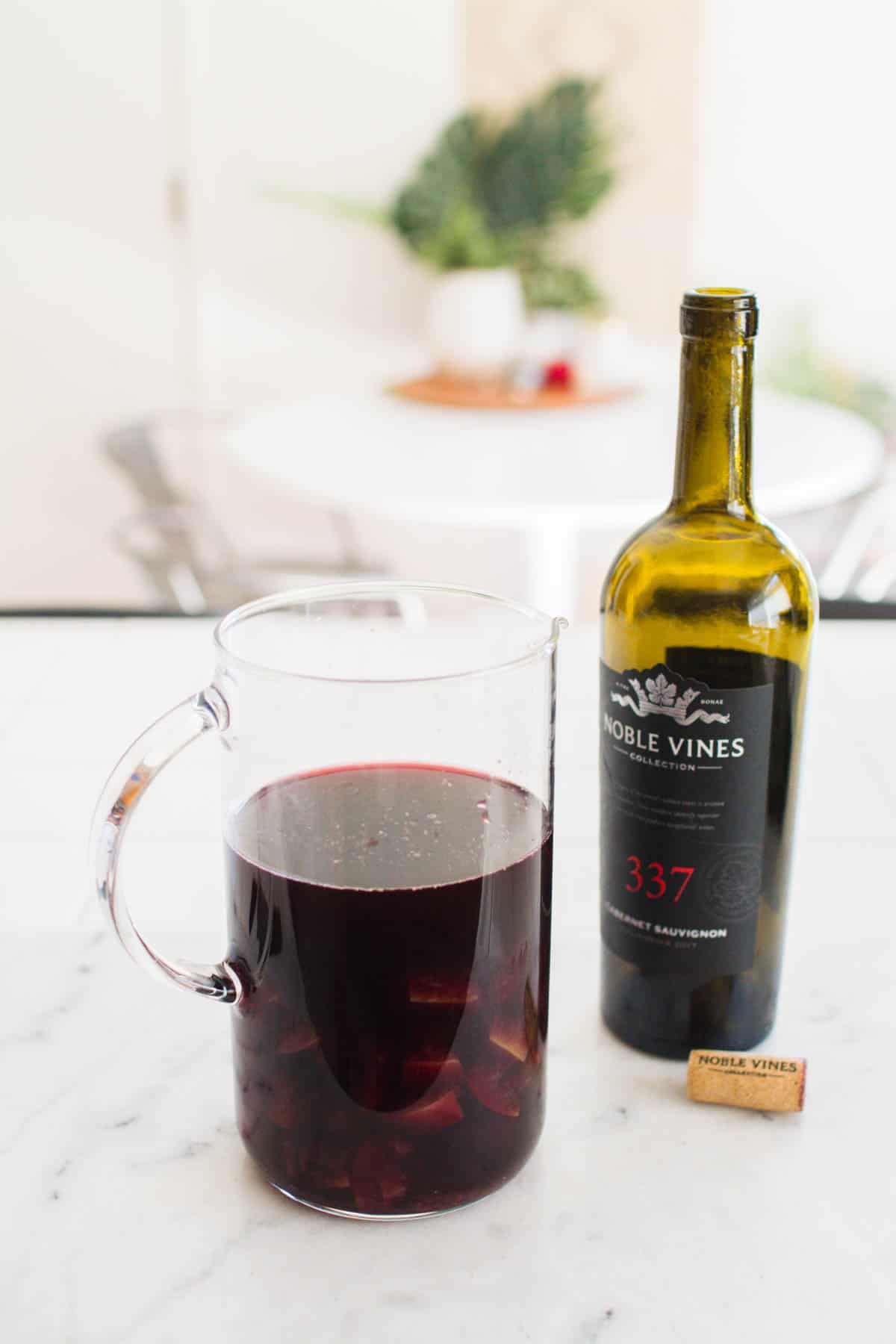 A pitcher of red wine and cooked apples next to an empty bottle of red wine.