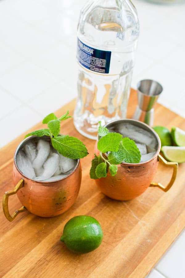 Copper mugs of mint garnished Moscow Mule cocktail on a wooden cutting board with a lime, jigger and bottle of vodka.
