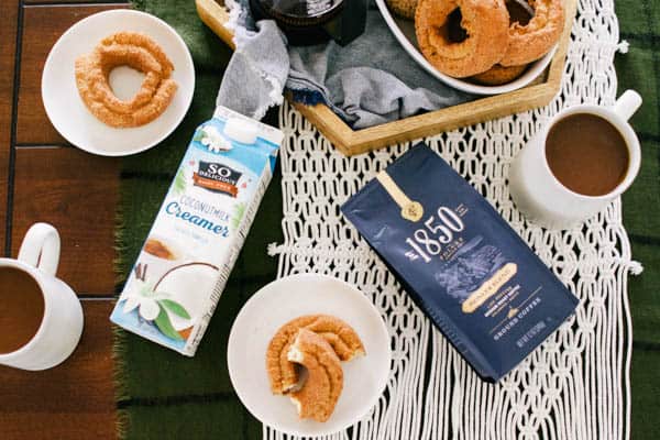 A package of coffee and creamer laying on a table next to cups of coffees next to a bowl of donuts.