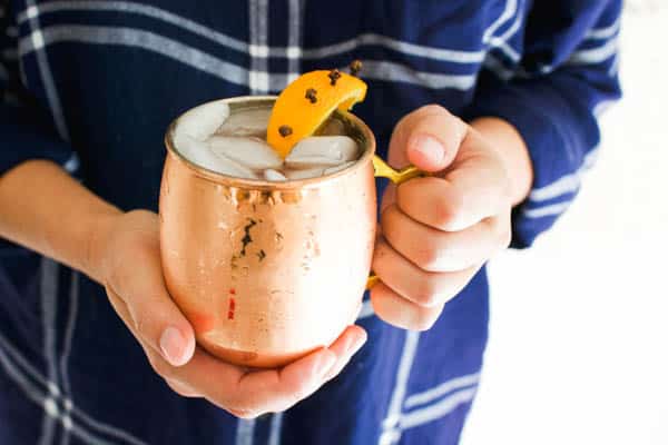 Woman holding a copper mule mug garnished with a clove studded piece of orange peel filled with a Holiday Moscow Mule cocktail.