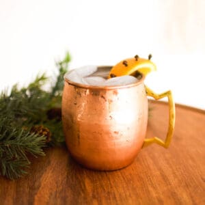 Close up image of a copper Moscow mule mug filled with a holiday themed mule and an orange slice garnish on a wood lazy susan.