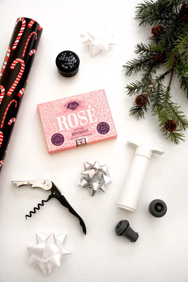 A box of rosé candies on a table next to a roll of holiday wrapping paper, pine sprigs, a silver bow, a wine key, and a wine bottle topper system. 
