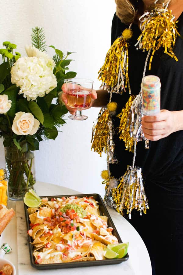 Woman holding wine and a confetti popper with metallic garden draped over her shoulders and a pan of lobster nachos on the table. 