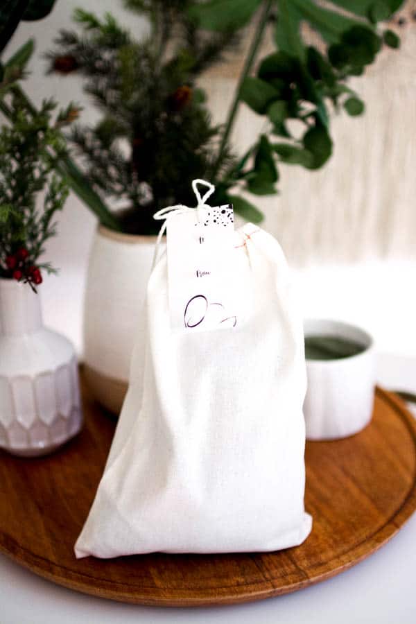 Small cloth gift bag on a wood lazy susan, surrounded by small white vases filled with greenery.
