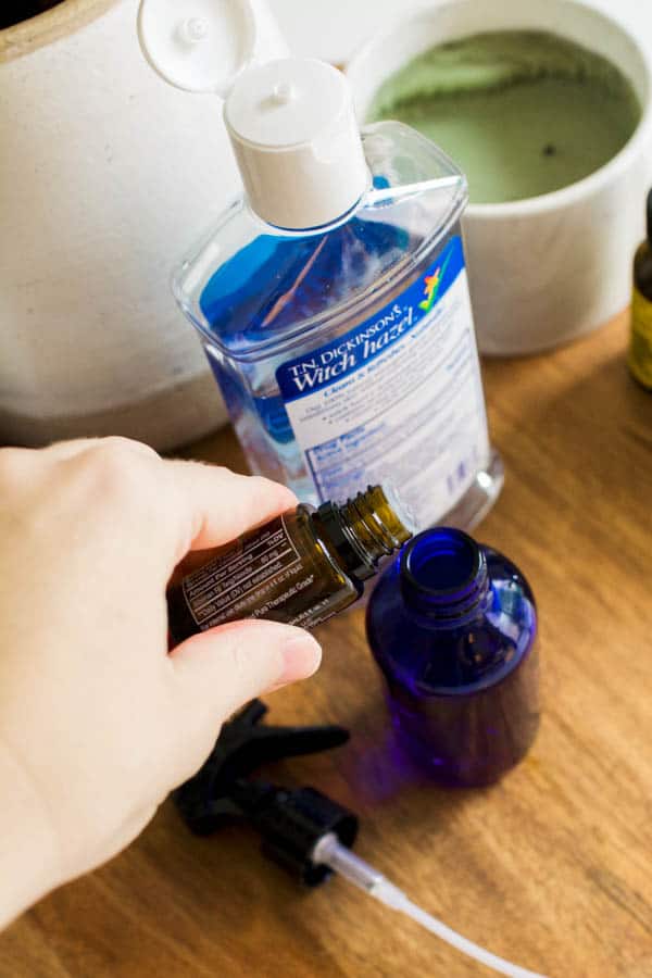 Adding essential oils to a blue glass bottle on a table next to a bottle of witch hazel. 