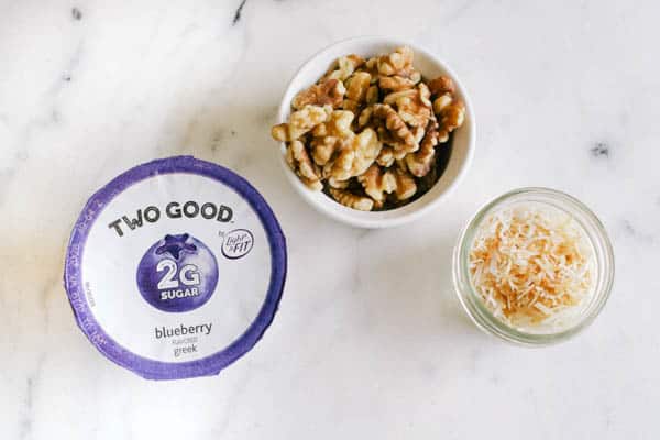 Overhead view of a yogurt container and small bowls of toasted coconut and walnuts.