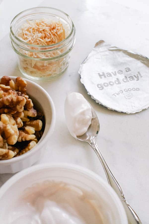 Yogurt on a spoon sitting on a table next to a bowl of walnuts, bowl of coconut and a yogurt lid that says "have a good day".