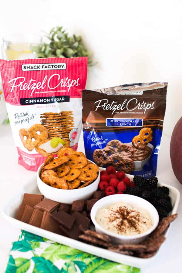 A bag of Cinnamon Sugar Pretzel Crisps next to a bag of Chocolate Covered Pretzel Crisps and a plate of berries, chocolate and a dessert dip in a bowl. 
