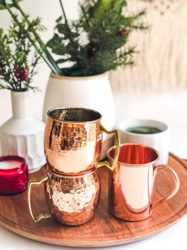 Copper mugs stacked on a wooden tray with candles and vases filled with greenery.