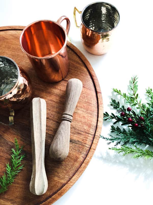 A wooden tray topped with muddling tools, copper mugs and greenery.
