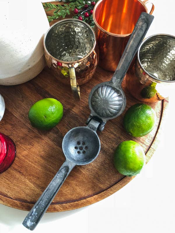 A wooden tray topped with copper mugs, fresh limes, and a citrus juicer.