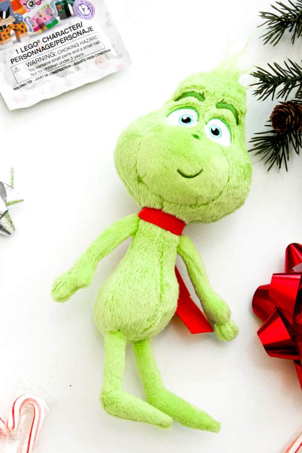 A stuffed Grinch doll on a white table next to some pine branches and a lego package. 