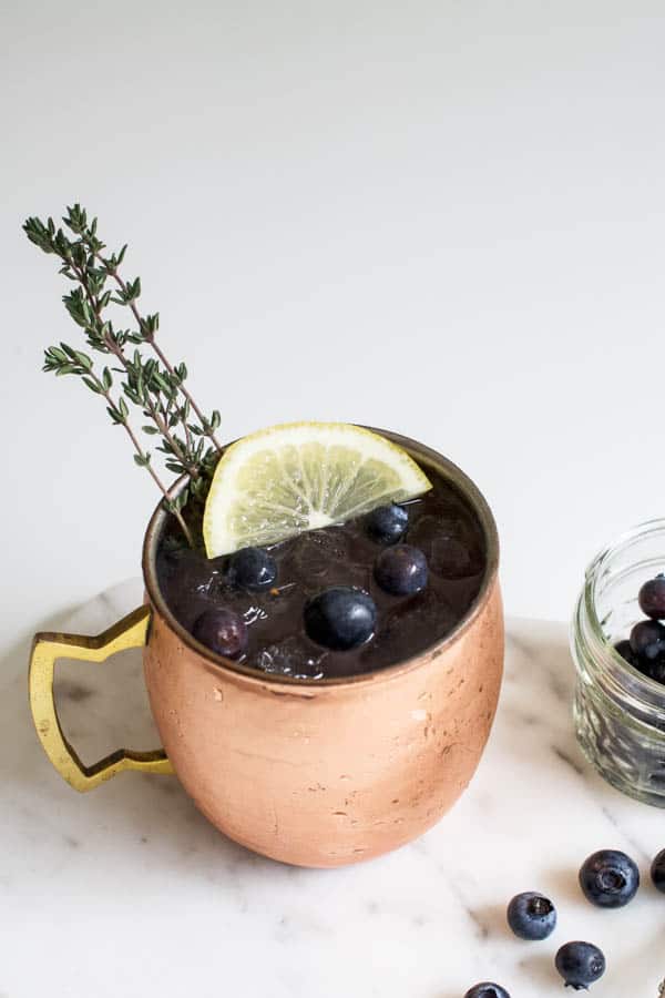 Blueberry Moscow Mule garnished with a lemon wedge and fresh thyme on a cutting board with fresh blueberries next to it.