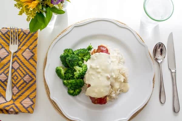 A white dinner plate with a creamy sauce topped chicken breast with rice and broccoli. 
