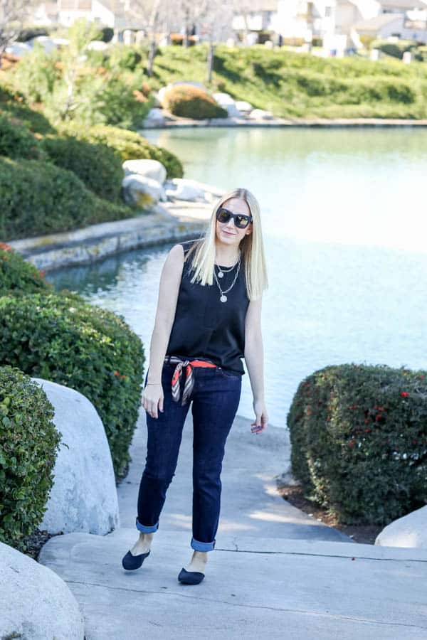 Woman in dark jeans with a black tank top and necklaces by a lake. 