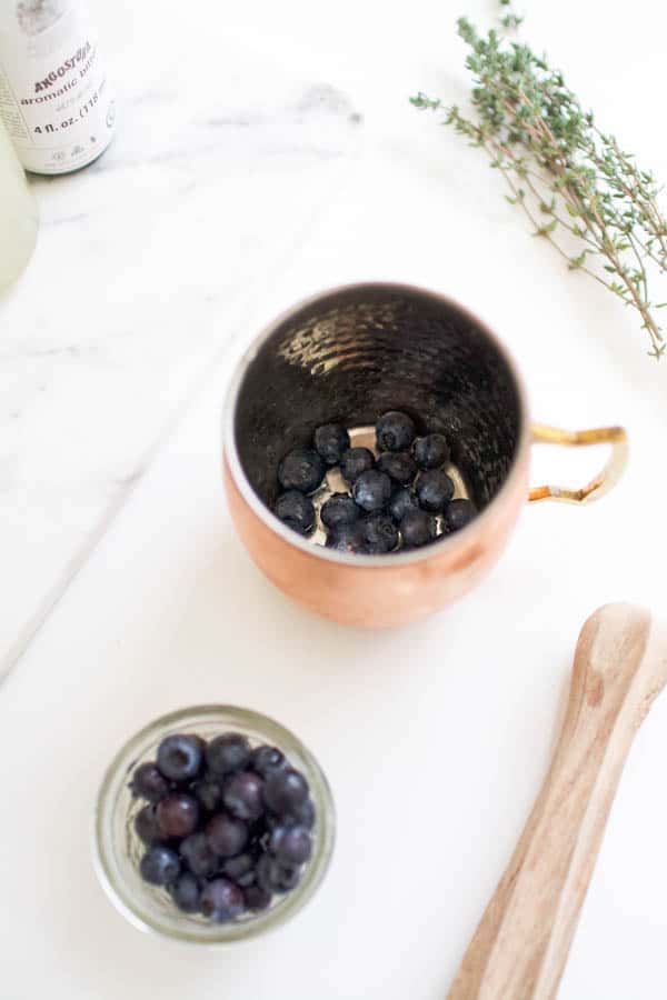 Fresh blueberries in the bottom of a copper Moscow Mule mug with a wooden muddler laying on the cutting board next to it.